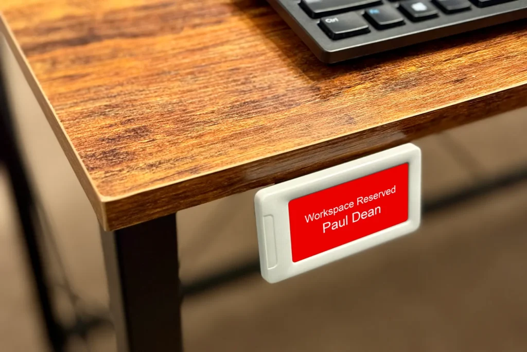 small white E Ink sign attached to the edge of a desk with red screen showing reservation