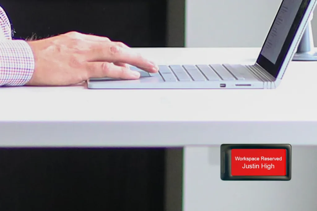 photo of man's hands working on laptop with e-paper desk sign attached to side of desk showing desk reservation