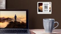 ePaper desk sign with desk reservation QR code in white block on graphic photo background mounted on wall behind desk with coffee mug and laptop