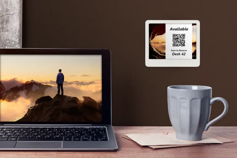 ePaper desk sign with desk reservation QR code in white block on graphic photo background mounted on wall behind desk with coffee mug and laptop