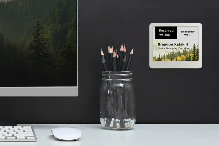 ePaper desk sign with desk reservation data on graphic background showing mountainside with trees mounted on wall behind desk with pencil jar, monitor, keyboard and mouse