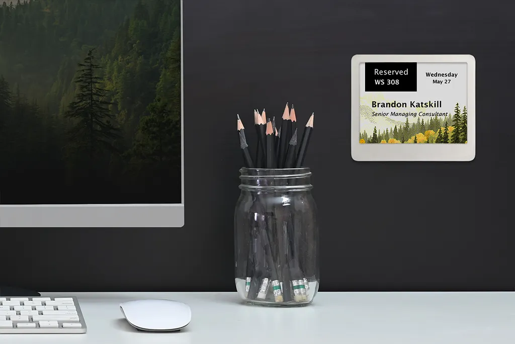 ePaper desk sign with desk reservation data on graphic background showing mountainside with trees mounted on wall behind desk with pencil jar, monitor, keyboard and mouse