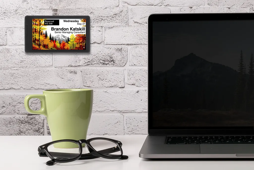 ePaper desk sign with colorful autumn tree graphic background showing desk reservation data mounted on wall behind a desk with a laptop, coffee mug and glasses