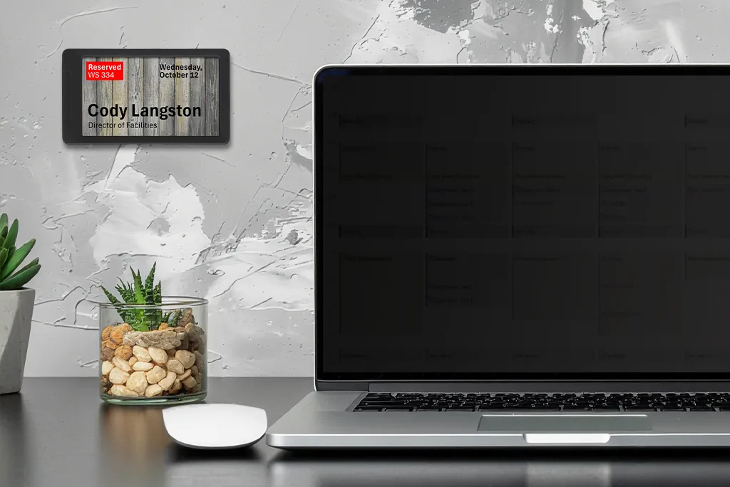 ePaper desk sign with wood panel background graphic showing current workspace reservation in red mounted on wall behind a desk with a laptop, mouse and plants