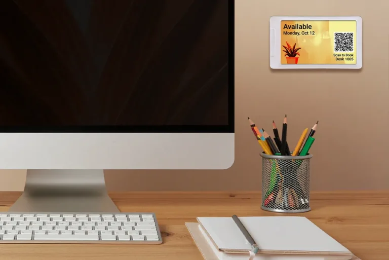 ePaper desk sign with plant background graphic and QR tag to make desk reservations mounted on wall beside monitor and above cup of pencils